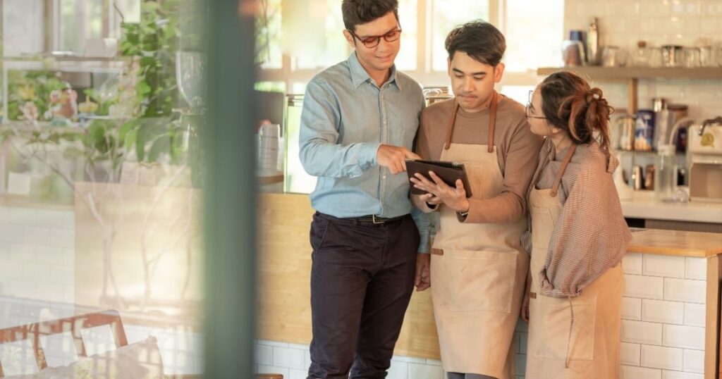 A manager or consultant pointing at a digital tablet while talking to two café staff members in aprons.