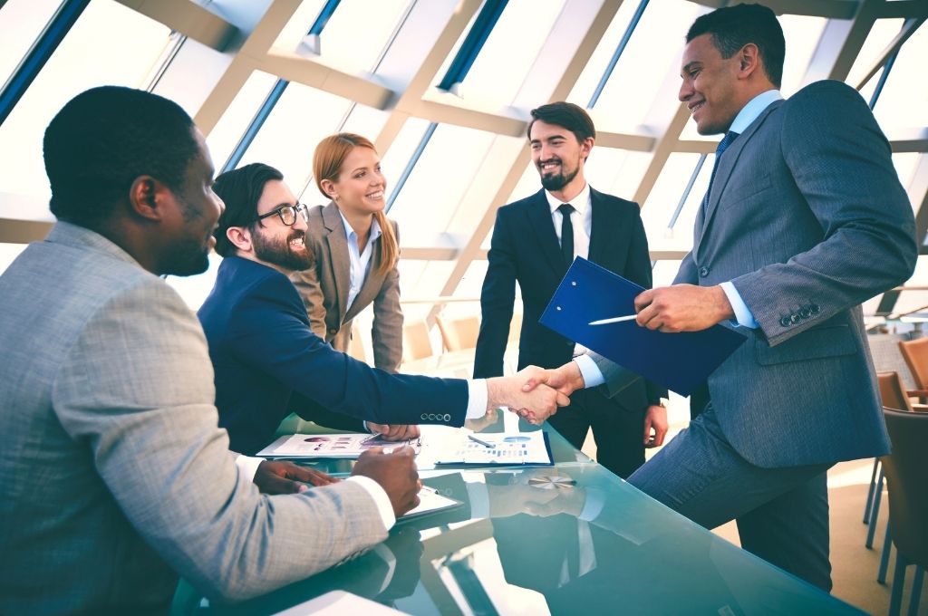 Diverse group of five businesspeople in a modern office, two men shaking hands to seal a deal.