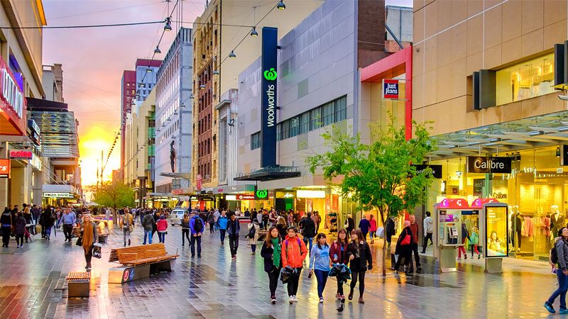 A crowded Woolworths-anchored pedestrian shopping street at sunset in an Australian city.