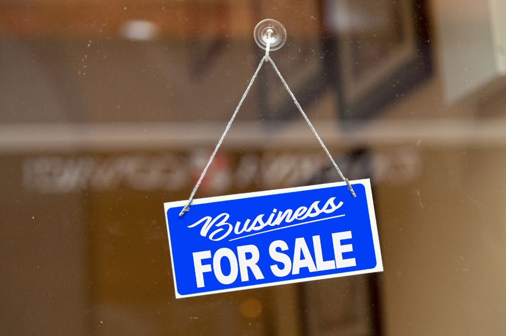 A bright blue sign with white script text that reads "Business FOR SALE" hanging on a glass door.